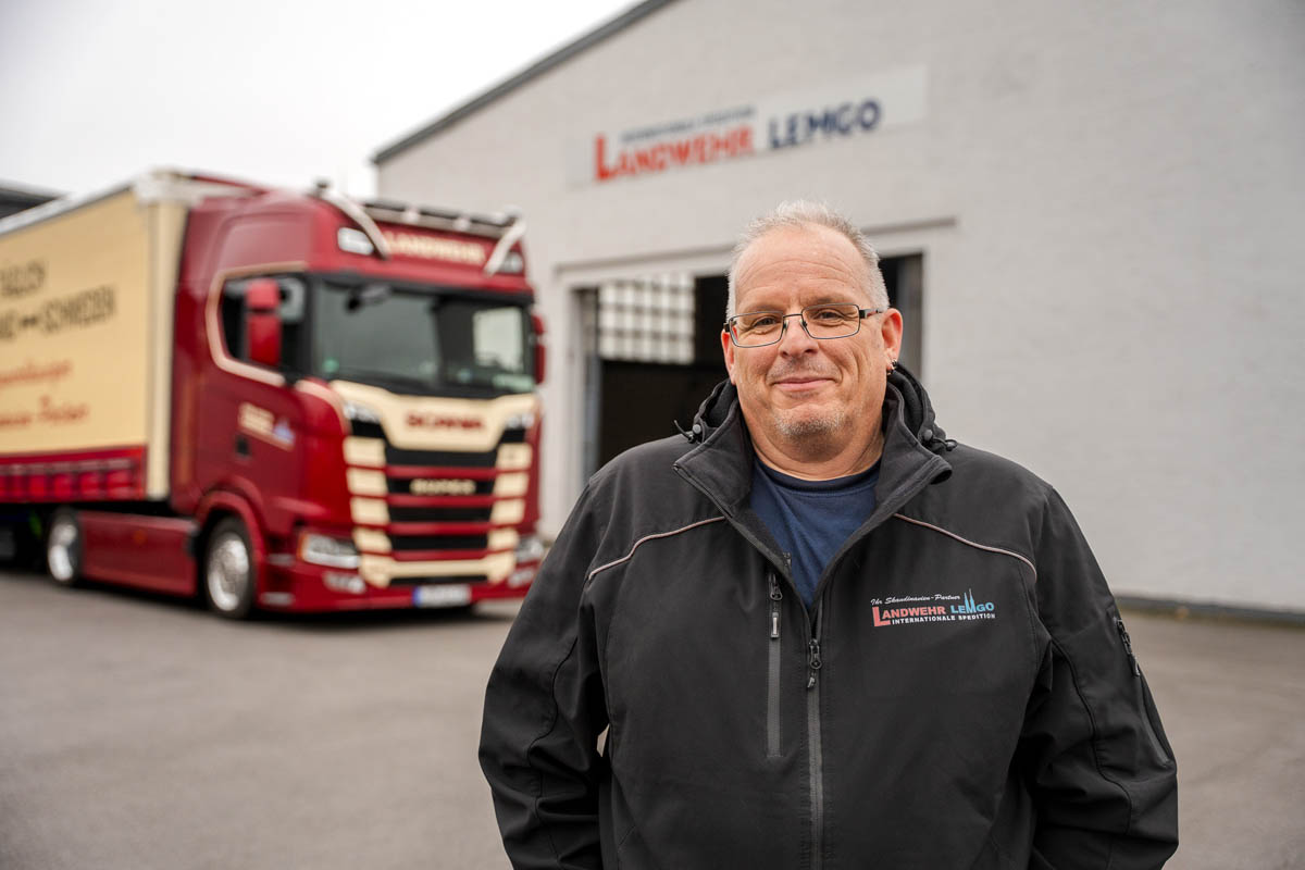 Ein Mann mit Brille und einer Jacke mit Landwehr Logo darauf steht sympathisch lächelnd vor einem roten LKW, der vor einer großen Lagerhalle geparkt ist.