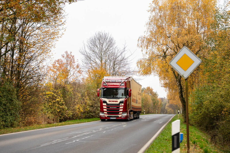Ein LKW der Spedition Landwehr fährt auf einer Landstraße, die von herbstlichen Bäumen umsäumt ist, auf die Kamera zu
