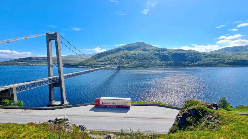 Blick von oben auf einen Fjord, an dem eine Landstraße verläuft. Ein Landwehr-LKW ist am Straßenrand geparkt.